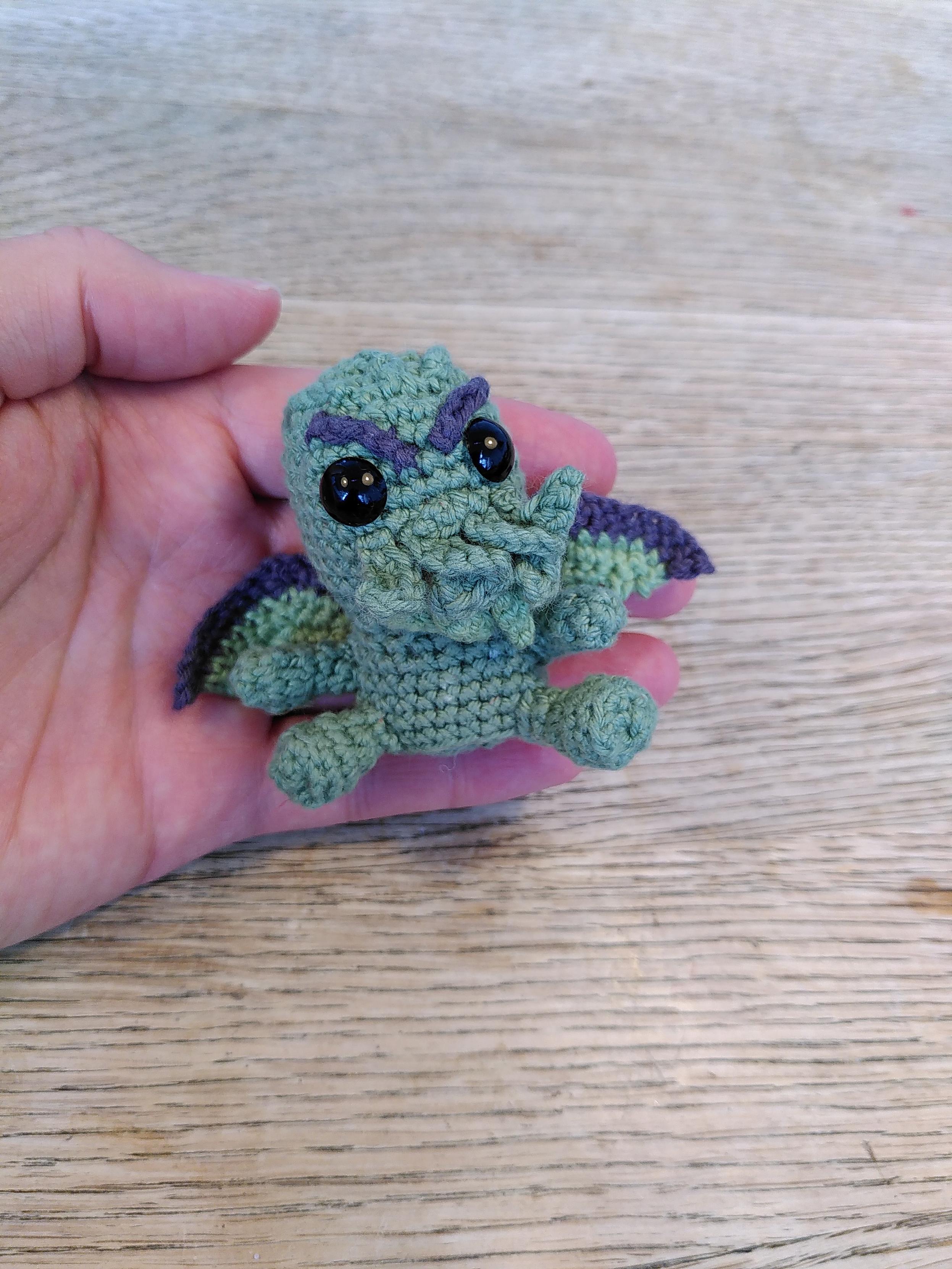 En hæklet minifigur af Chtulhu. Den er grøn, har tentakler i ansigtet og små vinger på ryggen. Den har store sorte øjne og øjenbryn der står på skrå så den ser godt sur ud. På billedet ligger den i min hånd så man kan se hvor lille den er. Dens lille størrelse gør den ret nuttet.